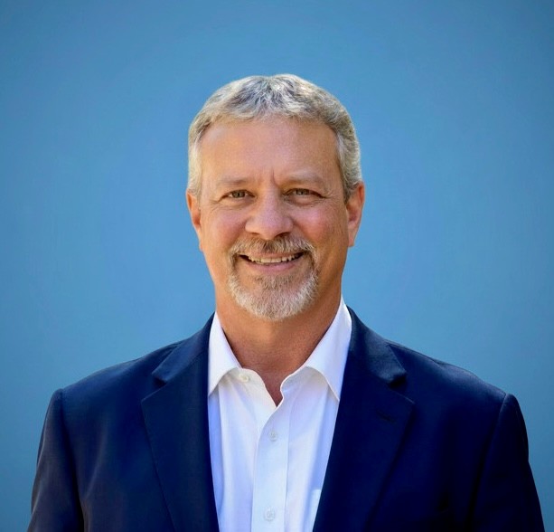 Professional headshot of a smiling middle-aged man in a navy suit and white shirt, posed against a blue gradient background.